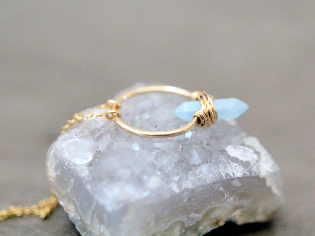 Crest necklace with a gold-toned hoop and pale blue aquamarine crystal resting on a light-colored stone.