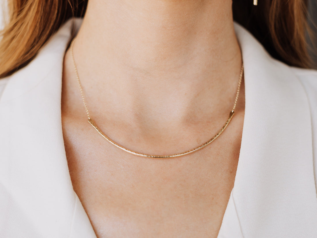 Model wearing the gold-toned Contour Collar Necklace with a curved textured bar and a white open-collar shirt.