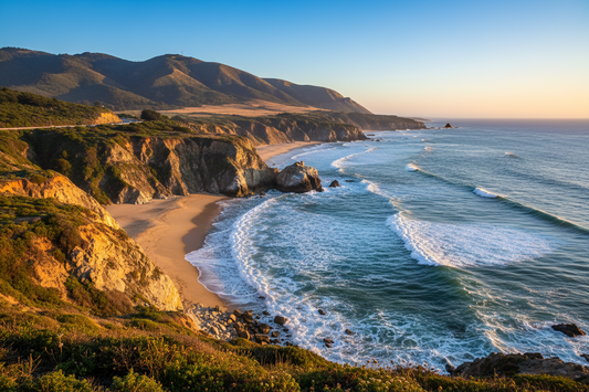 Golden coastal cliffs and sandy coves beside rolling ocean waves, with mountains in the background.