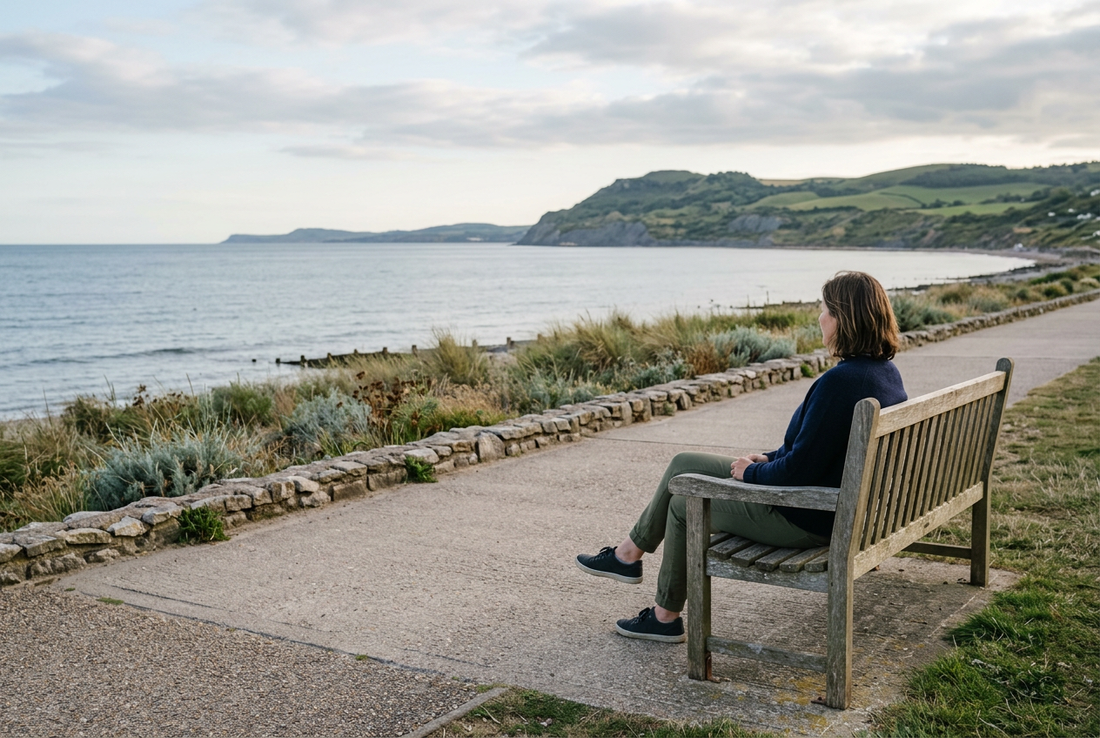 An adult seated on a wooden bench beside a coastal promenade, looking across an ocean bay toward green headlands.