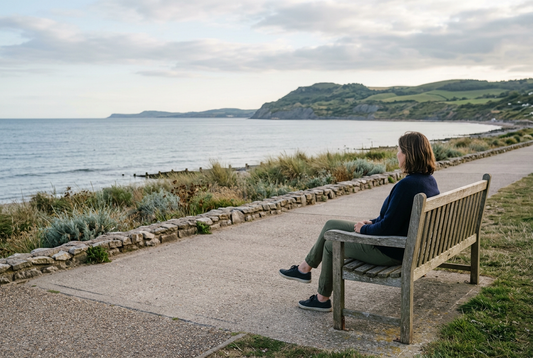 An adult seated on a wooden bench beside a coastal promenade, looking across an ocean bay toward green headlands.