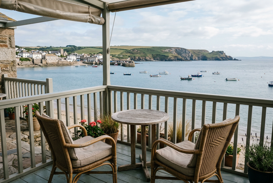 Two empty woven chairs and a small wooden table on a covered terrace overlooking boats in a bay, a coastal village, and green headlands.
